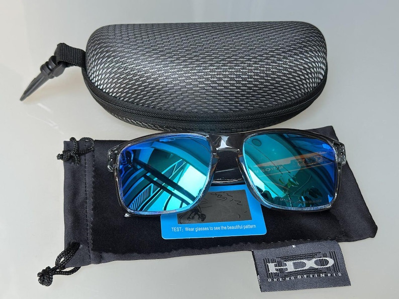 K&ouml;nnte beinhalten: Quadratische Sonnenbrille mit blau verspiegelten Gl&auml;sern, einem schwarzen Etui und einem schwarzen Beutel. Die Sonnenbrille hat einen transparenten Rahmen. Enth&auml;lt eine kleine blaue Karte und ein Etikett mit der Marke "HDO".