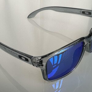 K&ouml;nnte beinhalten: Grau gerahmte Sonnenbrille mit blau reflektierenden Gl&auml;sern. Die Sonnenbrille hat eine rechteckige Form und ein schlankes, modernes Design. Die B&uuml;gel der Sonnenbrille sind leicht gebogen f&uuml;r einen bequemen Sitz. Die Gl&auml;ser reflektieren die Umgebung.