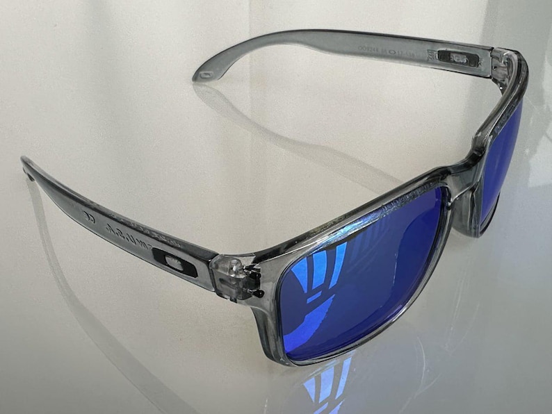K&ouml;nnte beinhalten: Grau gerahmte Sonnenbrille mit blau reflektierenden Gl&auml;sern. Die Sonnenbrille hat eine rechteckige Form und ein schlankes, modernes Design. Die B&uuml;gel der Sonnenbrille sind leicht gebogen f&uuml;r einen bequemen Sitz. Die Gl&auml;ser reflektieren die Umgebung.
