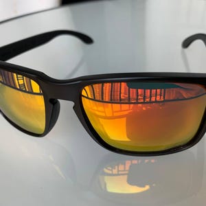 May include: Black sunglasses with orange-yellow lenses. The sunglasses have a rectangular frame with a subtle curve at the top and red accents on the temples. The lenses reflect the surrounding environment.