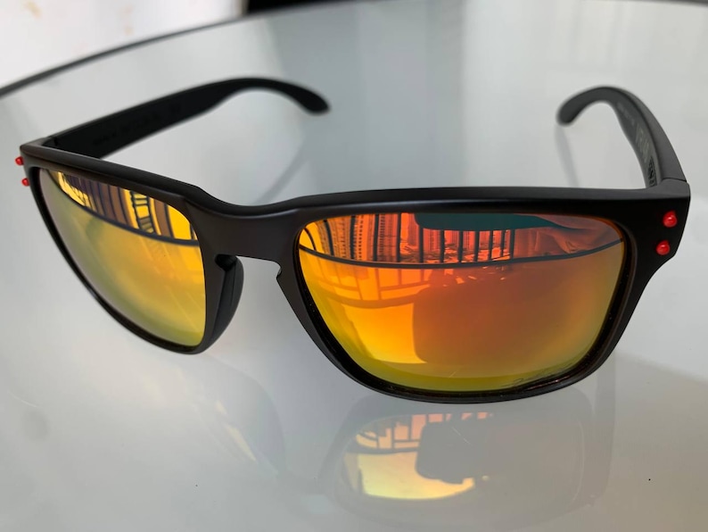 May include: Black sunglasses with orange-yellow lenses. The sunglasses have a rectangular frame with a subtle curve at the top and red accents on the temples. The lenses reflect the surrounding environment.