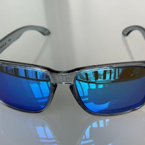 K&ouml;nnte beinhalten: Graue Sonnenbrille mit blau verspiegelten Gl&auml;sern. Die Sonnenbrille hat einen rechteckigen Rahmen und gebogene B&uuml;gel. Die Gl&auml;ser reflektieren die Umgebung und erzeugen einen lebendigen Blauton. Die Sonnenbrille ist f&uuml;r den Augenschutz konzipiert.