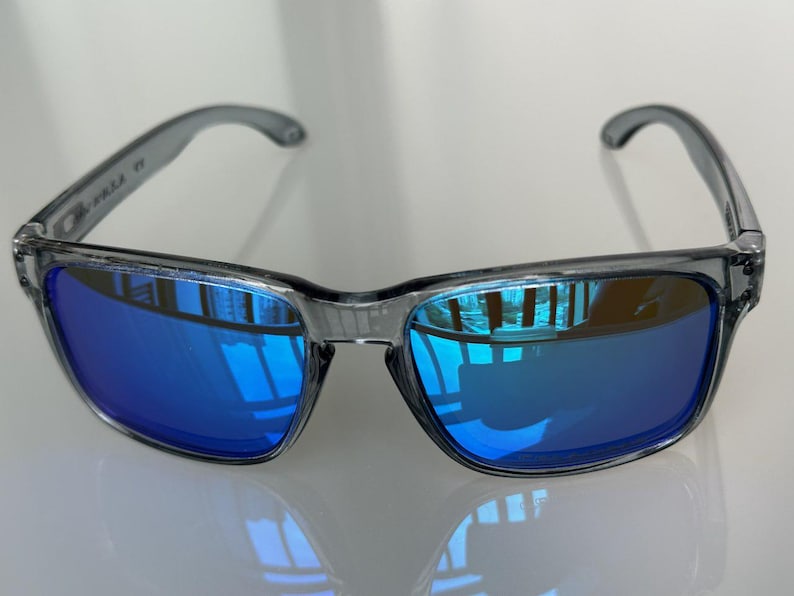 K&ouml;nnte beinhalten: Graue Sonnenbrille mit blau verspiegelten Gl&auml;sern. Die Sonnenbrille hat einen rechteckigen Rahmen und gebogene B&uuml;gel. Die Gl&auml;ser reflektieren die Umgebung und erzeugen einen lebendigen Blauton. Die Sonnenbrille ist f&uuml;r den Augenschutz konzipiert.