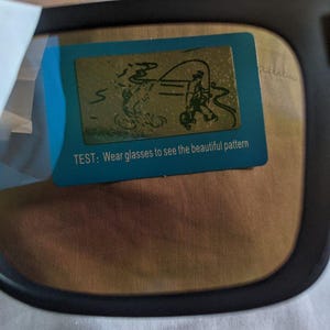 May include: Black-framed glasses with a blue and gold test card. The card features a drawing of a person fishing and the text: "TEST: Wear glasses to see the beautiful pattern."