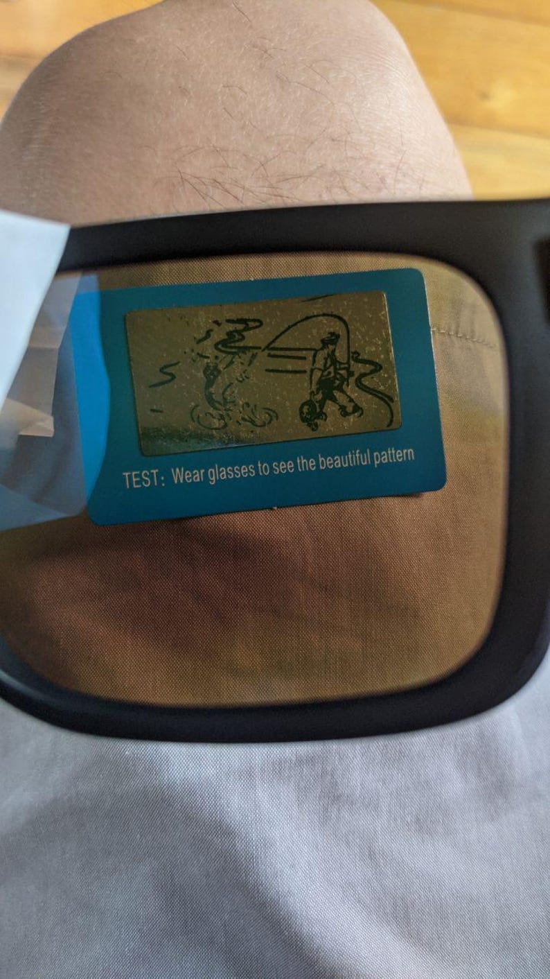 May include: Black-framed glasses with a blue and gold test card. The card features a drawing of a person fishing and the text: "TEST: Wear glasses to see the beautiful pattern."