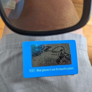 May include: Black-framed glasses with amber lenses are positioned above a blue card. The card has a silver design of a person fishing and the text: "TEST: Wear glasses to see the beautiful pattern."
