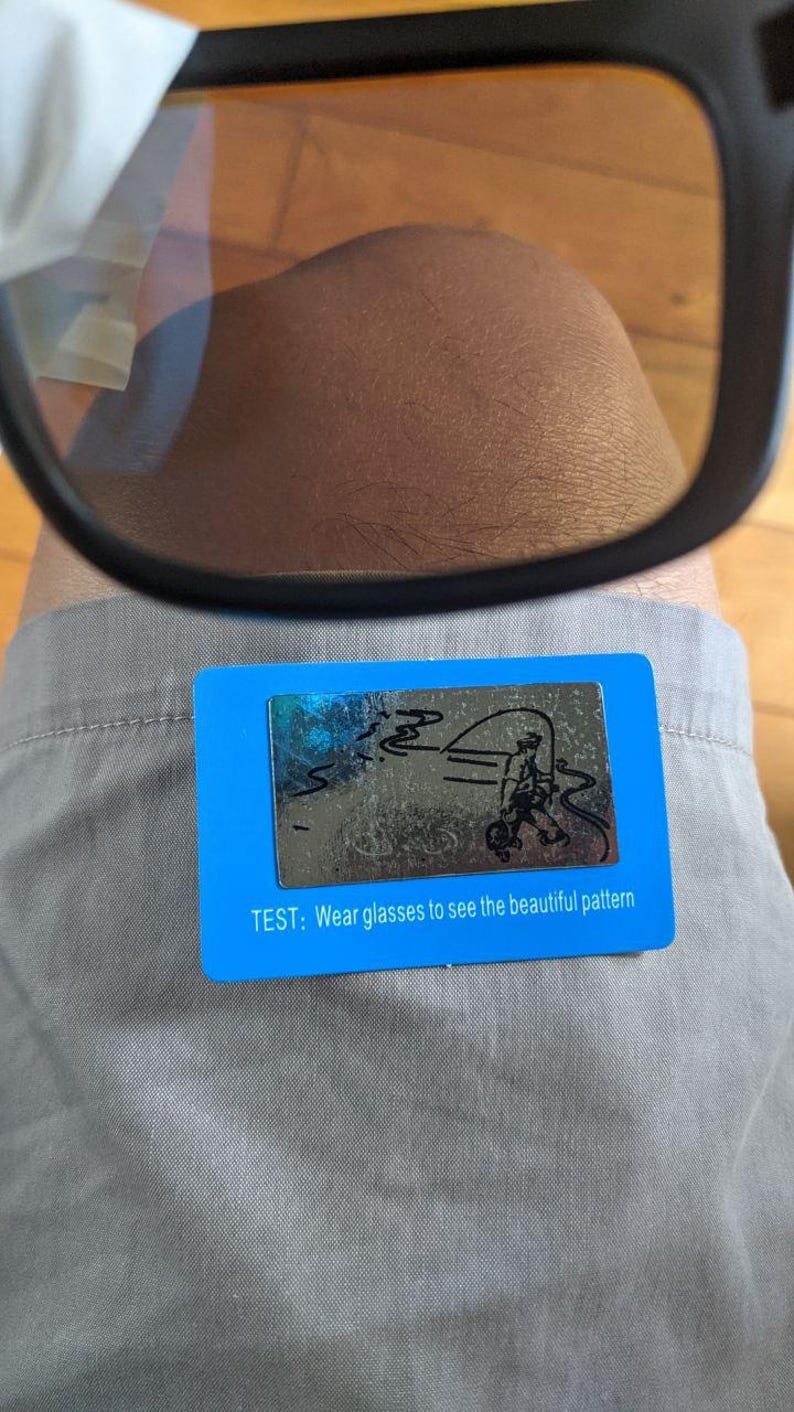 May include: Black-framed glasses with amber lenses are positioned above a blue card. The card has a silver design of a person fishing and the text: "TEST: Wear glasses to see the beautiful pattern."