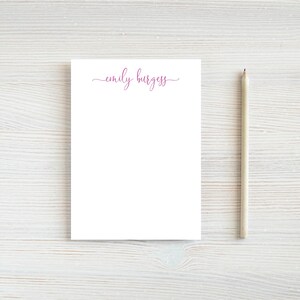 May include: A white notepad with a pink cursive inscription that reads "emily burgess" on the top. A wooden pencil is to the right of the notepad.