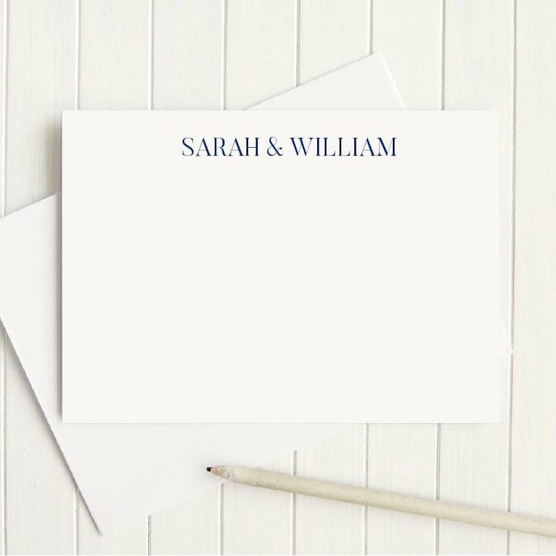 Op de afbeelding: Een witte kaart met de tekst "SARAH & WILLIAM" gedrukt in donkerblauwe inkt. De kaart ligt op een wit houten oppervlak met een potlood ernaast.