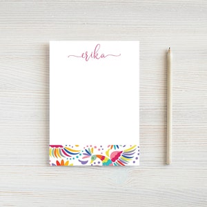 May include: White notepad with the name "Erika" in pink script at the top. The bottom of the notepad features a colorful floral design with birds. A wooden pencil rests on the right side of the notepad.