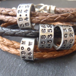 May include: Three braided leather bracelets in brown and black, each featuring a silver-toned metal ring charm. The metal rings are engraved with black numerical coordinates and geographic markers, including numbers and the letters N and W.