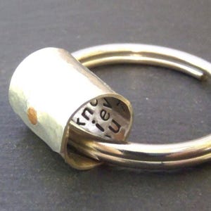 May include: A silver-toned ring featuring a cylindrical charm attached to the band. The exterior of the cylinder has a small gold-toned dot, while the interior is engraved with the visible black text that reads "know" and "iev" and "u".