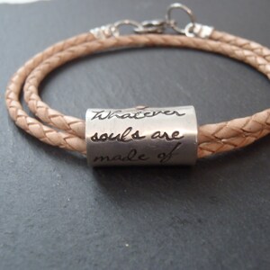 May include: A light tan braided leather cord bracelet featuring a cylindrical silver-toned metal bead. The bead is engraved with the cursive text "Whatever souls are made of". The bracelet includes a silver-toned metal clasp and circular ring.