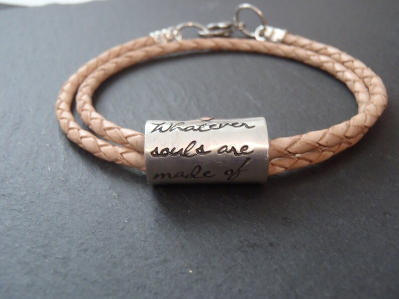 May include: A light tan braided leather cord bracelet featuring a cylindrical silver-toned metal bead. The bead is engraved with the cursive text "Whatever souls are made of". The bracelet includes a silver-toned metal clasp and circular ring.