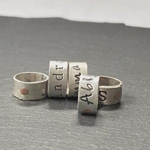 May include: Five silver rings with personalized names engraved on them. Each ring has a small, round, copper-colored accent. The names "J", "Andre", "Emma", "Abbi", and "S" are visible.
