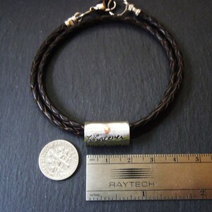 May include: A dark brown braided leather bracelet featuring a cylindrical silver-toned metal bead engraved with the word Whatever. The bracelet is shown next to a United States dime and a metal ruler measuring approximately 1.75 inches in length.