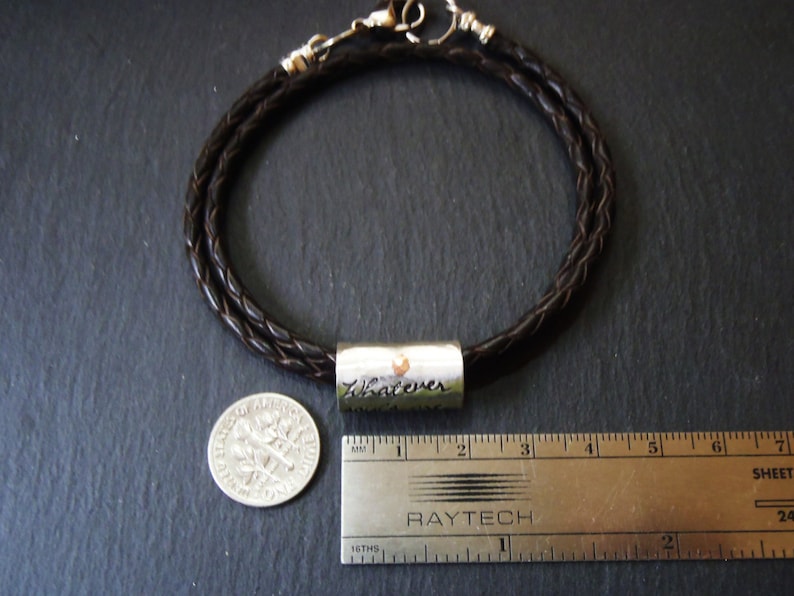 May include: A dark brown braided leather bracelet featuring a cylindrical silver-toned metal bead engraved with the word Whatever. The bracelet is shown next to a United States dime and a metal ruler measuring approximately 1.75 inches in length.
