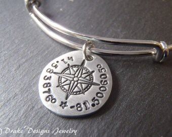 May include: A silver-toned expandable bangle bracelet featuring a round metal charm. The charm is stamped with a compass rose design in the center, surrounded by the numerical coordinates 41.583876 and -81.500605, along with a small star symbol.