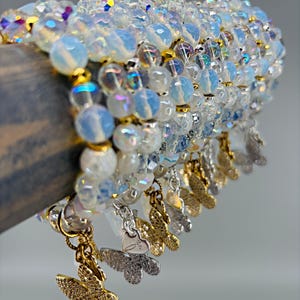 May include: A collection of beaded bracelets featuring iridescent, milky white round beads interspersed with small gold-toned spacers. Each bracelet is adorned with a dangling butterfly charm in either a gold or silver finish, adding detail.