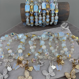 May include: A collection of beaded bracelets featuring iridescent white and clear glass beads, gold-toned spacers, and dangling butterfly charms. Some charms are solid white, while others are encrusted with small, sparkling clear crystals.