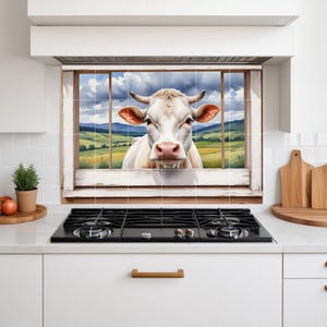 May include: A kitchen backsplash tile mural depicting a white cow peering through a window. The window reveals a rural landscape with green fields, hills, and a cloudy sky. The cow has brown ears and a pink nose.