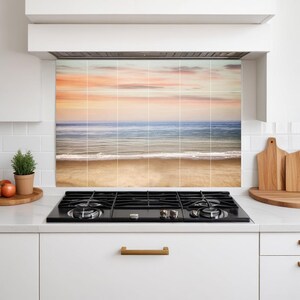 Op de afbeelding: Een keukenachterwand met een serene strandscène. De afbeelding toont een zandstrand, oceaangolven en een kleurrijke zonsondergang met tinten oranje, roze en blauw. De achterwand is geïnstalleerd boven een zwart kooktoestel.