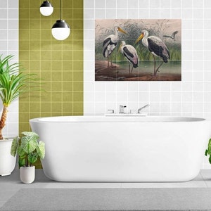 Puede incluir: Una bañera blanca con un grifo cromado y una pared de azulejos blancos detrás. Una planta verde está en una maceta blanca en el lado izquierdo de la imagen. Una planta verde está en una maceta blanca en el lado derecho de la imagen. Una impresión enmarcada de tres pájaros blancos con marcas negras de pie en un pantano está en la pared sobre la bañera.