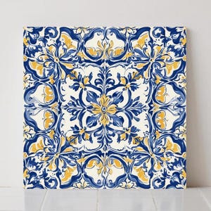 Puede incluir: Arte en lienzo cuadrado con un diseño floral simétrico. La obra de arte muestra un patrón repetitivo de flores y hojas azules y amarillas sobre un fondo blanco. El diseño recuerda a los azulejos tradicionales.