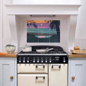 Puede incluir: Una cocina blanca con una parte superior negra y una pantalla digital de reloj. La cocina tiene seis quemadores y dos hornos. La cocina está en una cocina con un salpicadero de azulejos blancos del metro y una pintura de una puesta de sol sobre un lago.