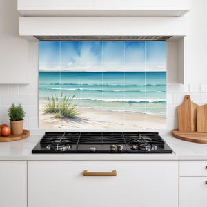 Op de afbeelding: Een keukenachterwand met een aquarel schilderij van een strandscène. Het kunstwerk toont een blauwe lucht, turquoise oceaangolven en een zandstrand met groene planten. De achterwand is geïnstalleerd boven een zwart fornuis.