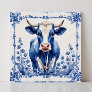 May include: A square tile with a blue and white cow illustration. The cow is depicted in shades of blue with a white face and pink nose. The tile is framed with a blue floral border and floral accents.