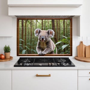 Op de afbeelding: Een keukenachterwand met een koala die uit een raam kijkt. De koala is grijs met pluizige oren en staat tegen een achtergrond van een weelderig groen bos. De achterwand is gemaakt van vierkante tegels.