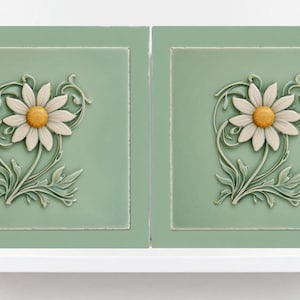 May include: Two square, sage green tiles featuring a raised daisy design. Each daisy has white petals and a yellow centre, surrounded by green leaves and vines. The tiles are displayed on a white shelf.