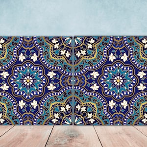 Puede incluir: Dos azulejos azules y dorados con intrincados diseños florales. Los azulejos tienen un patrón repetitivo de flores blancas, hojas azules y doradas y un diseño central en forma de estrella.