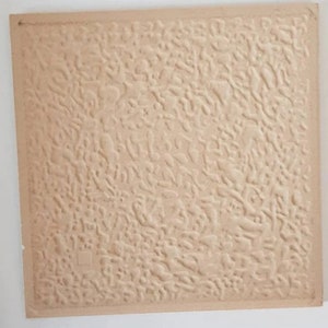 May include: A square, beige tile with a textured surface. The texture is a repeating pattern of raised, organic shapes, creating a tactile and visually interesting design. The tile's edges are slightly darker than the center, adding depth.