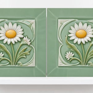 May include: Two green ceramic tiles featuring a raised white daisy design. The daisies have yellow centres and are surrounded by green leaves.