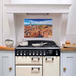 Può includere: Una cucina bianca con una cucina a gas nera e crema. Il paraschizzi è piastrellato e presenta una fotografia panoramica di uno skyline urbano. La fotografia è di una città con tetti in tegole rosse e un alto campanile.