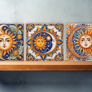 Puede incluir: Tres baldosas de cerámica cuadradas con diseños de sol y luna. Cada baldosa presenta una cara central de sol o luna con intrincados patrones florales y celestiales en naranja, amarillo y azul. Las baldosas se exhiben en una estantería de madera.