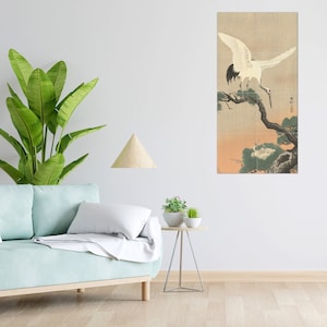 May include: A white crane with black wing tips perched on a branch of a pine tree. The crane is looking down at a smaller crane on the ground. The background is a soft, peachy-orange colour.