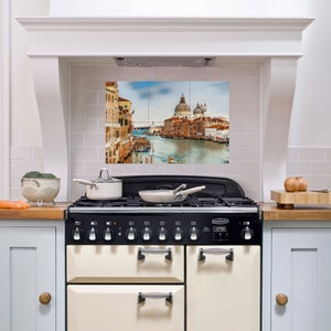 May include: A white kitchen with a black and cream range stove. The backsplash is white tile with a picture of a canal in Venice, Italy.