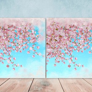 May include: Two watercolor paintings of pink cherry blossoms hanging over a blue sky. The paintings are framed and displayed on a wooden floor.