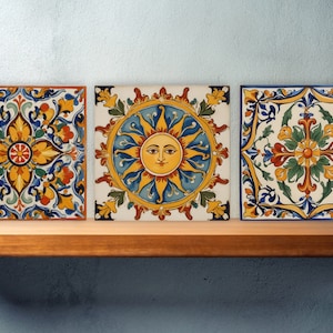 May include: Three square ceramic tiles with intricate floral designs. The center tile features a yellow sun with a human face. The tiles are painted in shades of blue, yellow, red, and green.