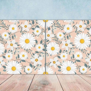 May include: A seamless pattern featuring white daisies with yellow centres on a light pink background. The daisies are surrounded by green leaves and small white flowers.