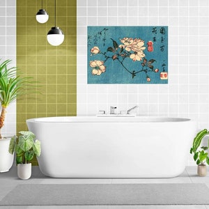 Puede incluir: Una impresión floral japonesa enmarcada con flores color crema y hojas verdes sobre un fondo verde azulado. La obra de arte cuelga sobre una bañera blanca en un baño. La impresión incluye caracteres japoneses.