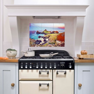 May include: A white kitchen with a black and cream range stove. The backsplash is white tile with a colorful landscape image of a rocky coastline with a small building.