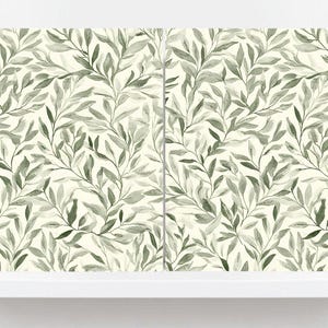 Könnte beinhalten: Ein Tapetendesign mit einem sich wiederholenden Muster aus olivgrünen Blättern und Zweigen vor einem cremefarbenen Hintergrund. Die Blätter haben ein weiches, verschwommenes Aussehen und erzeugen eine natürliche Ästhetik. Die Tapete wird auf einem weißen Regal präsentiert.