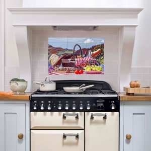 May include: A kitchen backsplash with a printed image of a picnic scene. The image features a wicker basket, a red and white checkered tablecloth, a bottle of wine, two glasses of wine, grapes, and tomatoes. The backsplash is installed above a white and black range.