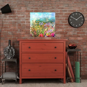 Op de afbeelding: Een rode houten dressoir met drie lades en zwarte knoppen. Het dressoir staat voor een bakstenen muur met een schilderij van een kleurrijk bloemenveld en een zwarte klok aan de muur.