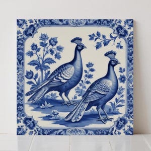 May include: A square ceramic tile featuring a blue and white peacock design. Two peacocks are depicted with detailed plumage and crests, surrounded by floral and foliate patterns. The tile has a decorative border with similar floral motifs.