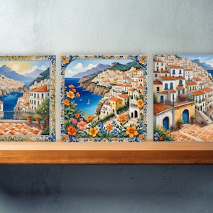 May include: Three framed paintings depicting a colorful Italian coastal town with white buildings, blue water, and green hills. The paintings are framed in a decorative blue and yellow border with floral accents.